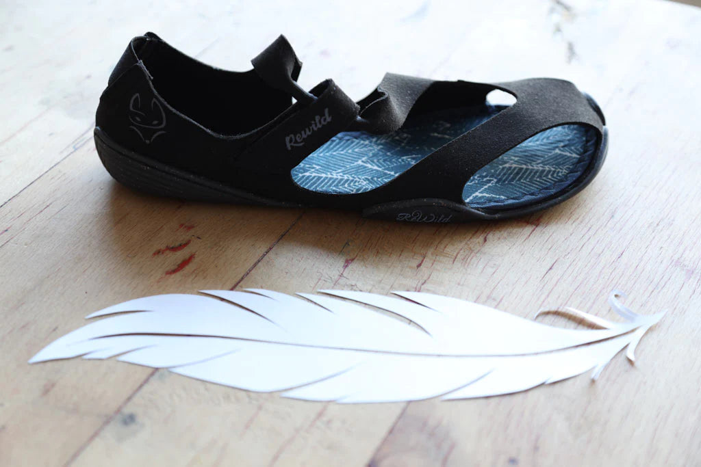 A black Wildling Shoes sandal on wooden floorboards, a large white feather next to it.