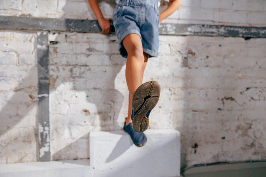 In front of a light-colored brickwork, a person in short jeans and Wildling minimal shoes stands on a low, light cuboid. The person is in the frame up to the hips and stretches one leg forward so that the sole of one shoe is turned towards the camera.