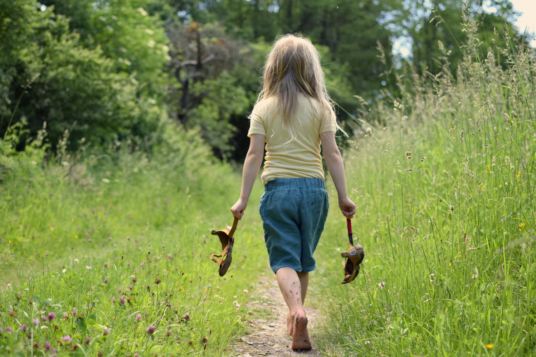 A young person with long blond hair, T-shirt and shorts walks barefoot, back turned to the camera, on a narrow path through a flowering meadow, carrying Wildling Shoes sandals in their hands. 