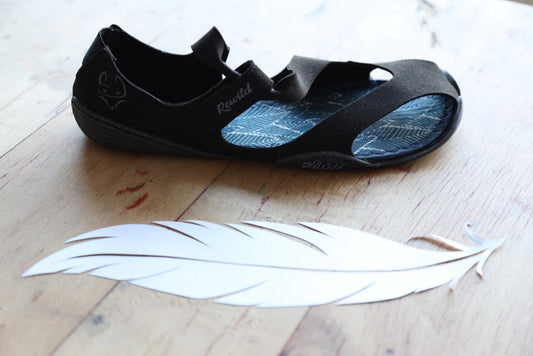 A black Wildling Shoes sandal on wooden floorboards, a large white feather next to it.