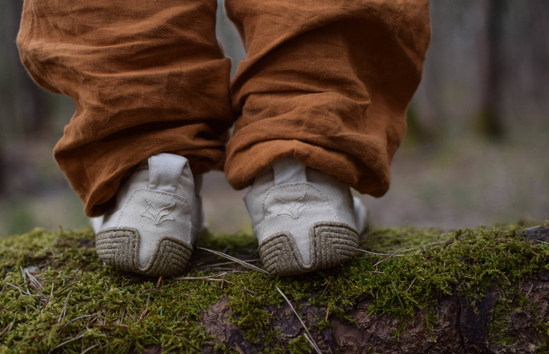  Close-up of two feet to mid-calf from behind. They are standing on a lying, mossy tree trunk and are wearing Wildling barefoot shoes and ocher-colored long trousers.