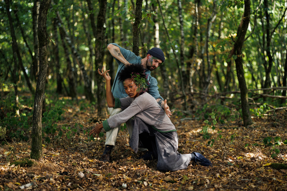 Two people are gently intertwined as they dance. Both are wearing autumn apparel and an ornament of green foliage on their heads. There are trees around them, and the ground is covered with foliage. Poetic, autumnal mood.