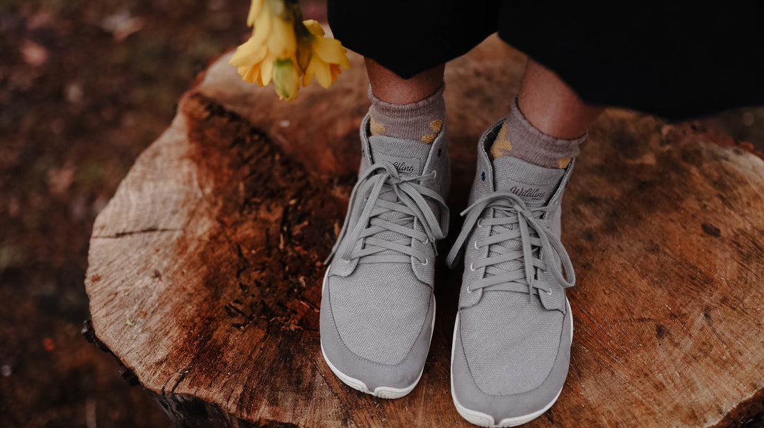 Close-up: Two feet in grey Wildling shoes standing on a tree stump. Yellow flowers at the edge of the picture.