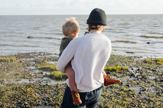 An adult holding a child in their arms, photographed from behind. The person is standing on a stony beach and looking out to sea.
