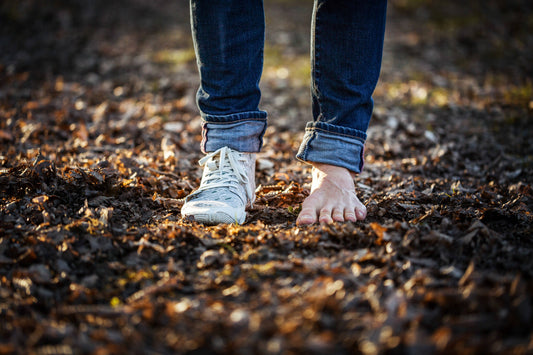 Close-up of two feet on leaf-covered ground. One of the feet is wearing a Wildling minimal shoe, the other foot is barefoot.